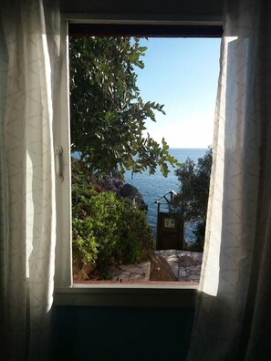 A window with a view of the sea and rocky coastline, curtains partially drawn, and a small structure with a sign.