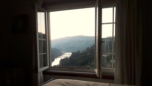 Een kamer met een bed, een raam met gordijnen en uitzicht op een rivier en heuvels.
