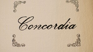De tekst “Concordia” in een decoratief lettertype, omgeven door versierde hoekornamenten.