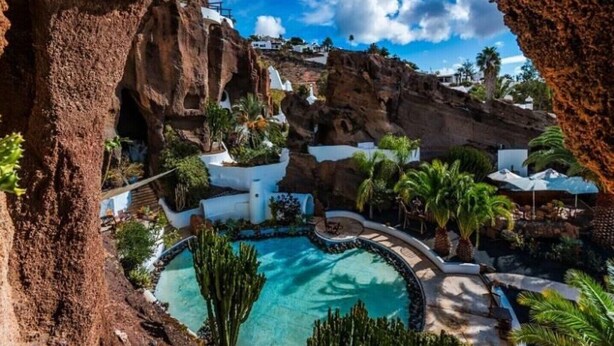 Un complexe hôtelier doté d’une piscine, entouré de bâtiments blancs et de palmiers, le tout se dressant sur un fond de falaises rocheuses et sous un ciel bleu azur.