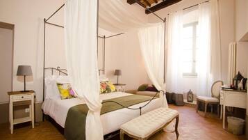 A four-poster bed with white drapes, a bedside table with a lamp, a bench, and a small desk with a chair.
