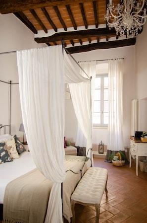 Een slaapkamer met een hemelbed, een bankje, een kroonluchter en een raam met gordijnen.