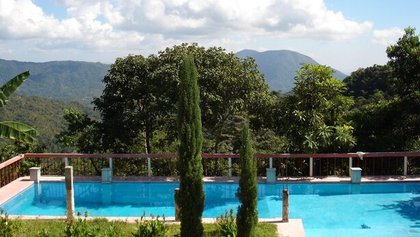 A rectangular swimming pool with a clear blue water surface, surrounded by a red deck and white pillars. In the background, there are lush green trees and a mountain range under a partly cloudy sky. - Eco Hotel El Refugio, In the mountains but near the sea. - ROOM 8 (PLUMA HIDALGO)