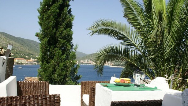 Un espacio de comedor frente al mar con mobiliario de mimbre, una mesa con un mantel individual verde y vistas al mar y las colinas.