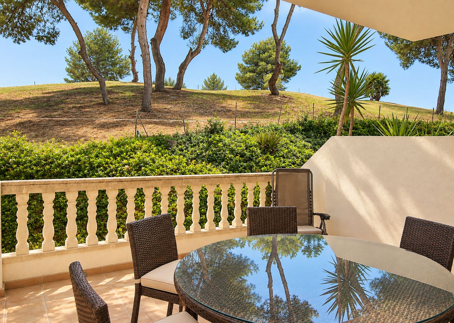 An outdoor patio with a round glass table, wicker chairs, and a view of a landscaped hill with trees and shrubs.