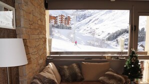 Een gezellige woonkamer met een stenen muur, een groot raam met uitzicht op een besneeuwde berg, een kleine kerstboom en een moderne bank met zachte kussens.