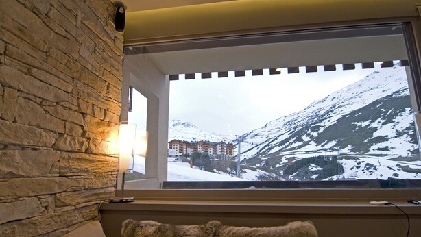 Een kamer met een stenen muur, een raam met uitzicht op besneeuwde bergen en gebouwen, en een bont deken op de bank.