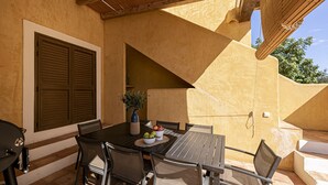 Une terrasse extérieure avec une table dressée pour le repas, entourée de chaises, et un barbecue.