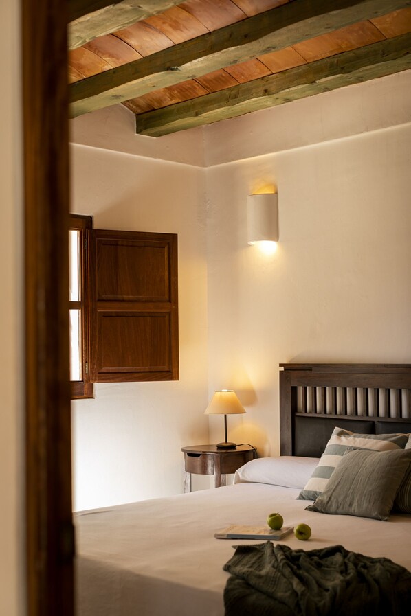 A bedroom with a wooden ceiling, a bedside lamp, a bed with pillows, and a nightstand with a book and apples.