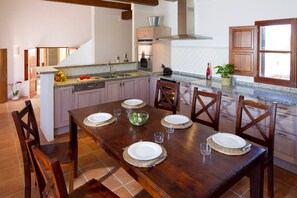 Un comedor con una mesa de madera dispuesta para seis personas, rodeada de sillas de madera. Se ve una cocina con electrodomésticos modernos y una ventana al fondo.