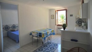 Una habitación pequeña y luminosa con una cama, una mesa con mantel, una ventana con vista a palmeras y un rincón de kitchenette.