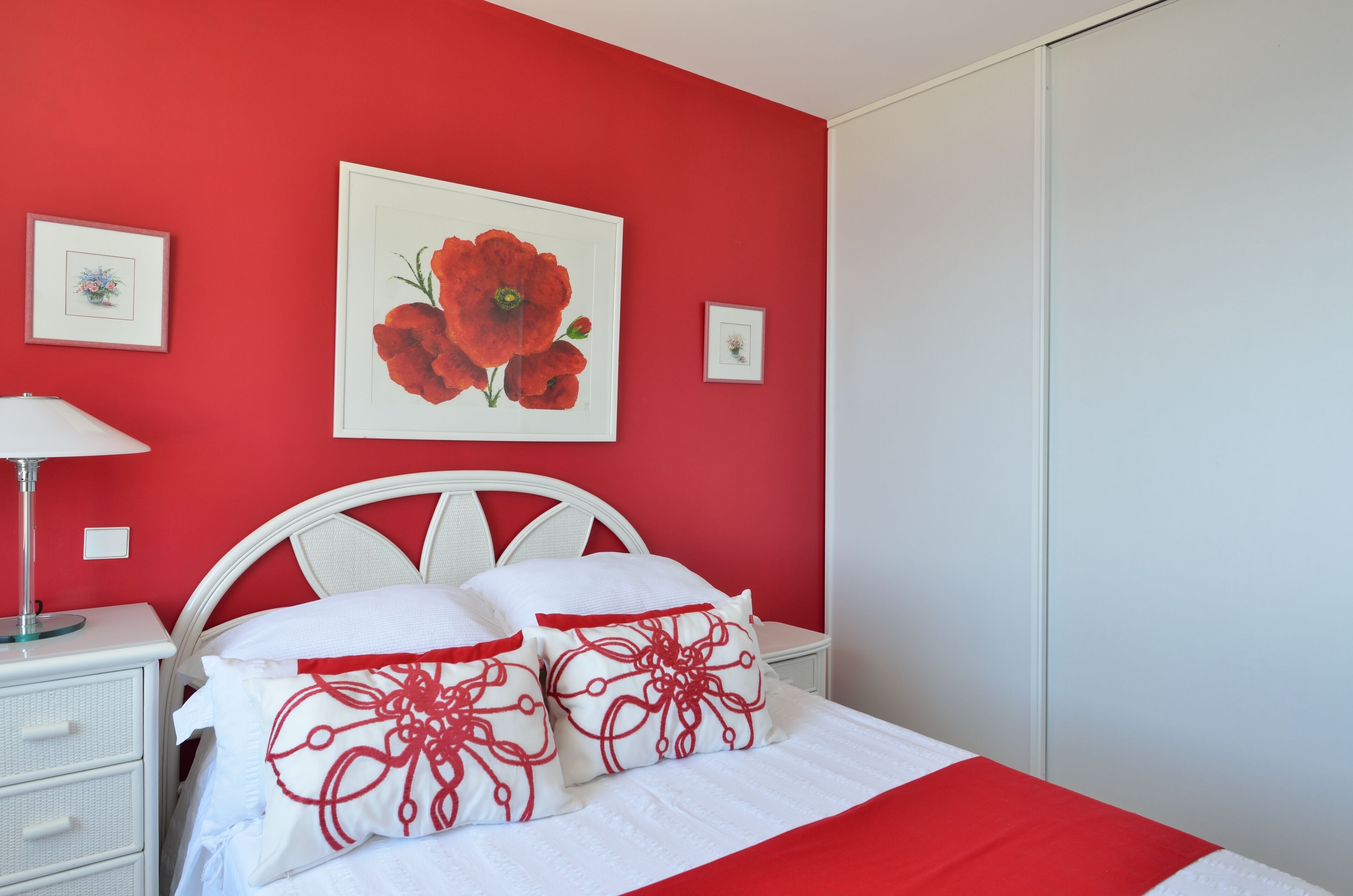 A bedroom with a red wall, white bed, and a framed flower painting.
