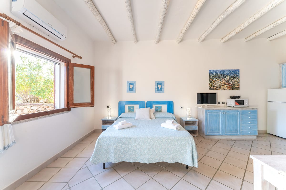 Een slaapkamer met een bed, een raam, een schilderij, een televisie en een kitchenette.