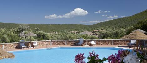 A swimming pool with lounge chairs and umbrellas, surrounded by greenery and hills.