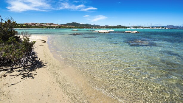 En strand med klart vand, en sandet kystlinje og et fjerntliggende syn af bygninger og både.