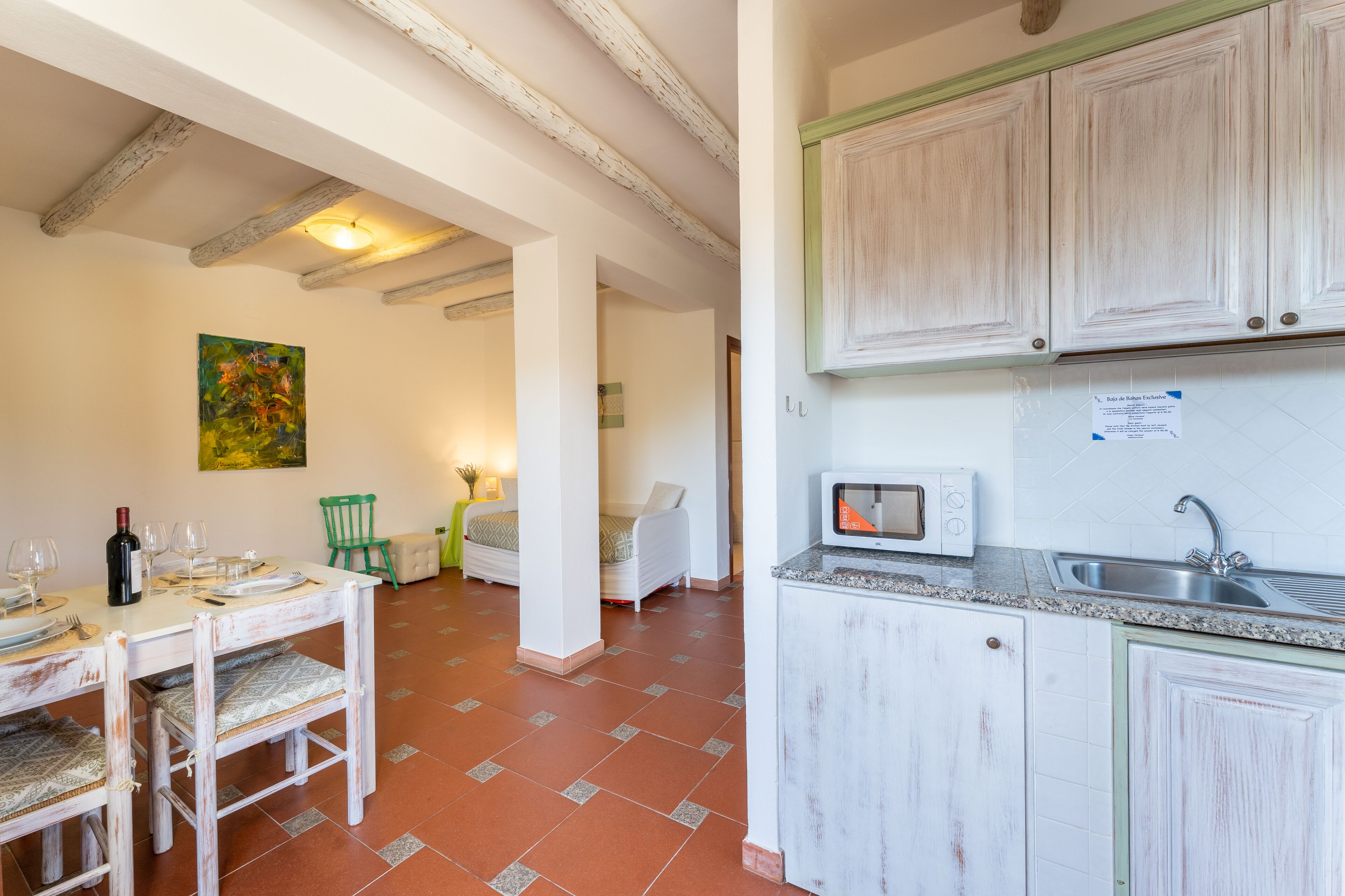 A kitchen with white cabinets, a microwave, and a sink. There is a dining table with chairs and a painting on the wall.