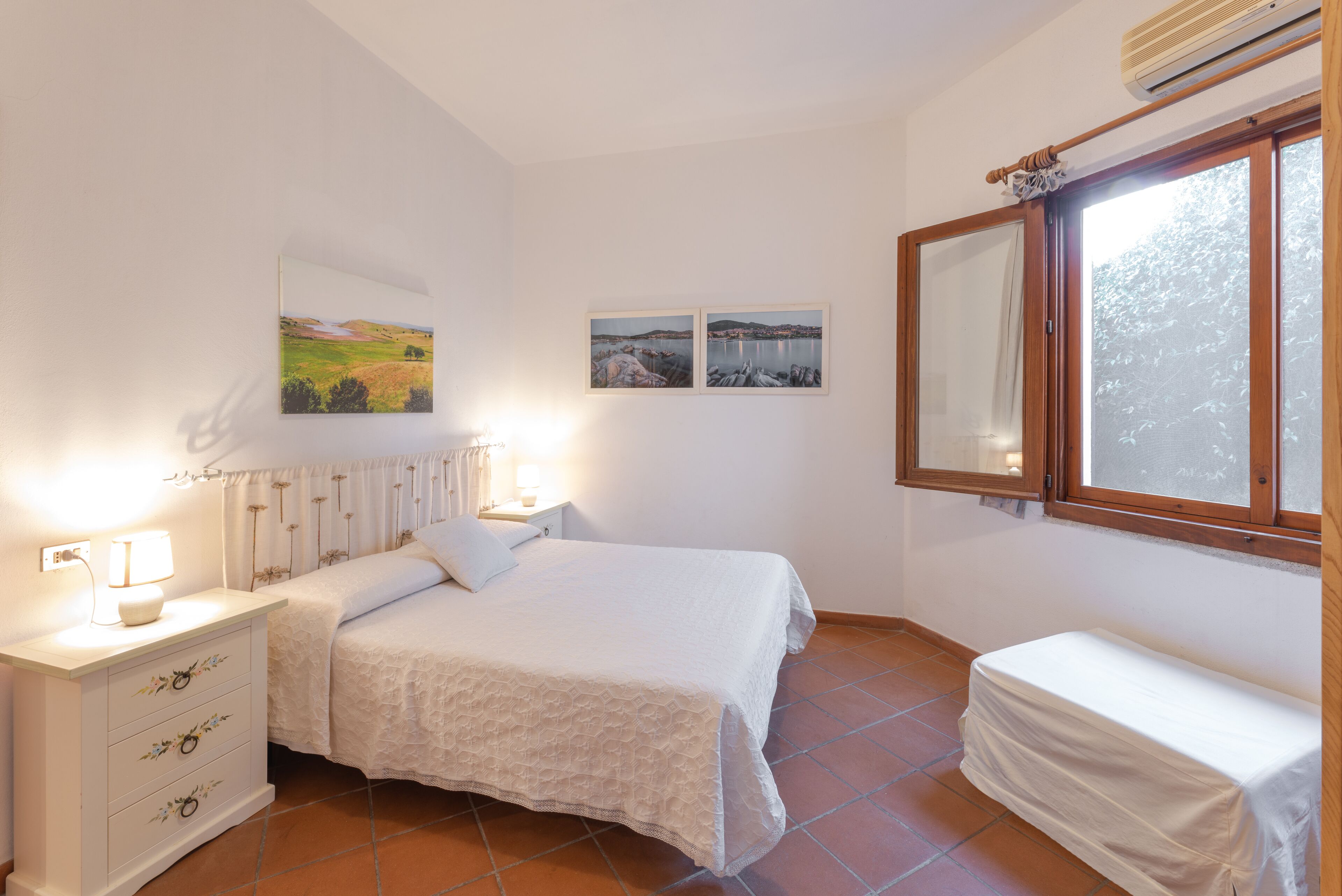 A bedroom with a bed, a bedside table with a lamp, a small bedside table with a lamp, a window with a view of trees, and a wall with two framed pictures.