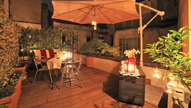 Un patio exterior con una mesa de comedor, sillas y un paraguas grande. Hay plantas en macetas y velas alrededor de la mesa.