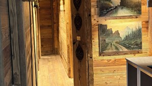 A wooden interior hallway with a wooden ceiling, a wall-mounted light fixture, and framed landscape paintings.