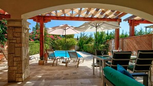 Un espacio exterior techado con piscina, sillas de descanso y una mesa con sombrillas.