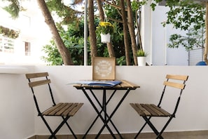 A small outdoor seating area with a table and two chairs, surrounded by greenery and a white wall. - Urban Beach Sand (1Y) (Rethymno)