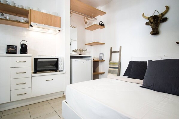 A compact bedroom with a bed, a small kitchenette, and a wooden shelf unit.