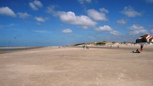 Ein weiter, sandiger Strand mit Fußspuren, einigen Personen und einem Gebäude mit rotem Dach im Hintergrund.
