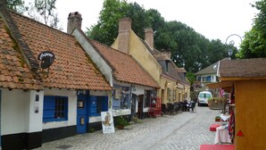 Eine Kopfsteinpflasterstraße gesäumt von traditionellen Gebäuden, einige mit blauen Fensterläden, andere mit Ziegelöfen. Es gibt Außenbereiche mit Sitzgelegenheiten und eine kleine Holzkonstruktion mit rotem Tischleinentuch.