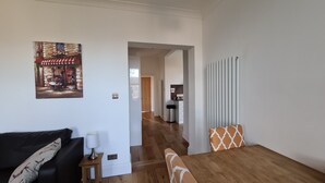 A modern living room with a wooden floor, a black sofa, a dining table with orange cushions, and a painting of a rustic scene. - Large sunny fresh two bedroom apt in London ideal for tourists, refurbished  (London)