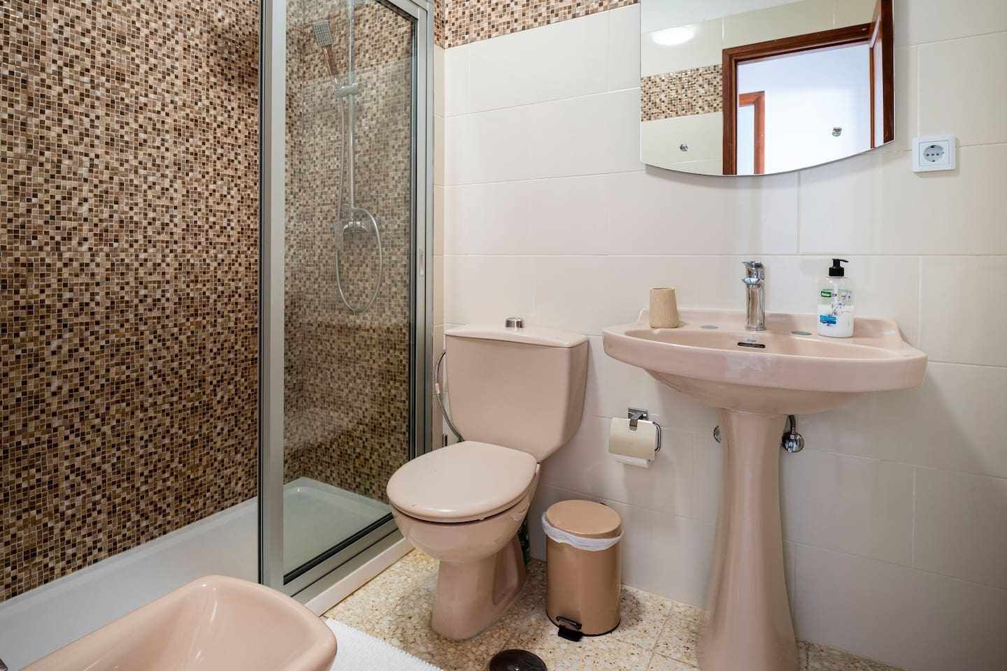 A bathroom with a glass-enclosed shower, a toilet, a sink, and a trash bin.