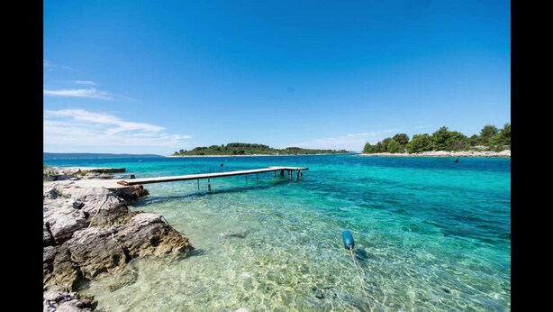 Una vista costera despejada con un muelle que se adentra en aguas turquesas, costa rocosa y una isla distante cubierta de vegetación.