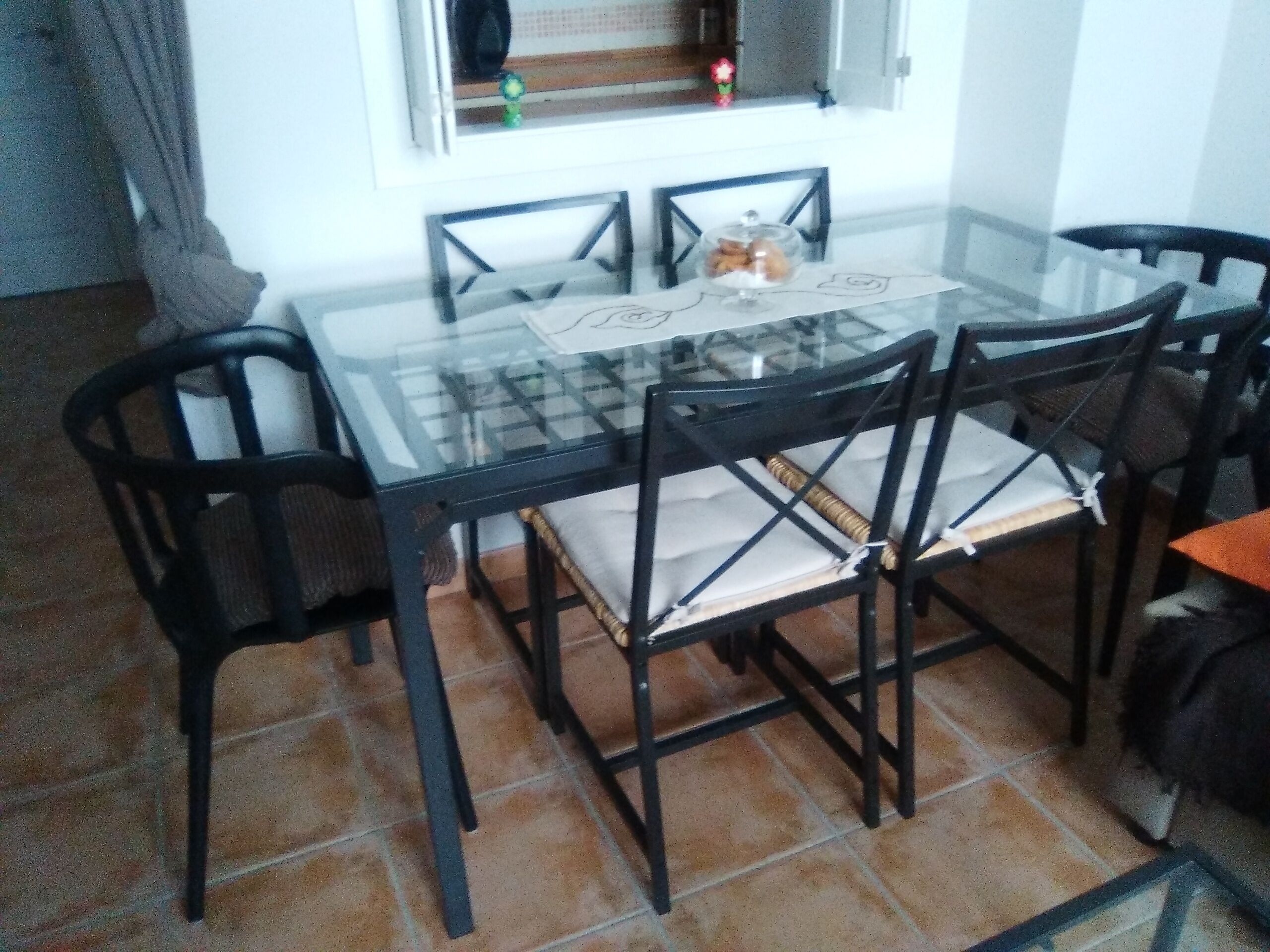 Una mesa de comedor con cubierta de vidrio, cuatro sillas, una ventana con vistas y piso de baldosas.