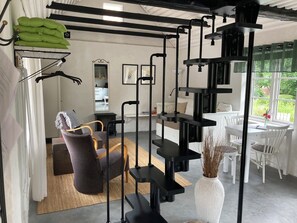 A room with a black exercise station, a white dining table with chairs, a grey sofa, and a view of greenery outside. - Charming cottage on a farm (Ystad V)