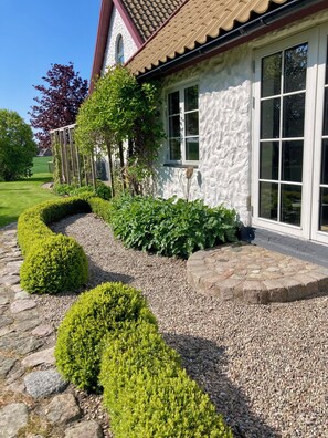A house with a stone pathway leading to a white door, surrounded by well-maintained hedges and a garden. - Charming cottage on a farm (Ystad V)