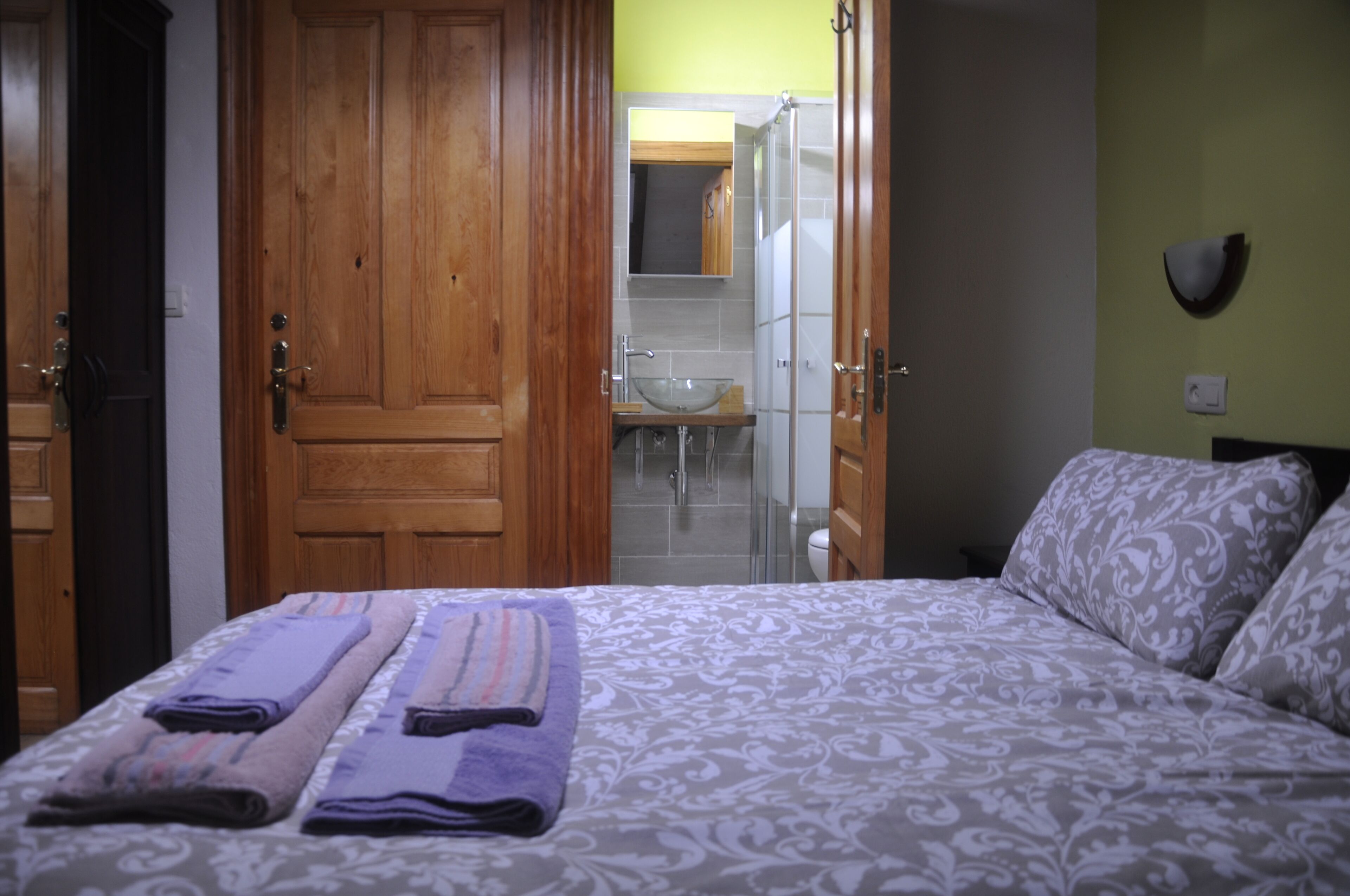 Habitación de hotel con cama, puerta de madera, baño con lavamanos y espejo, y ducha.