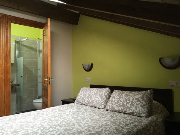 A bedroom with a bed, a nightstand, a wall-mounted light fixture, and a bathroom visible through an open door.