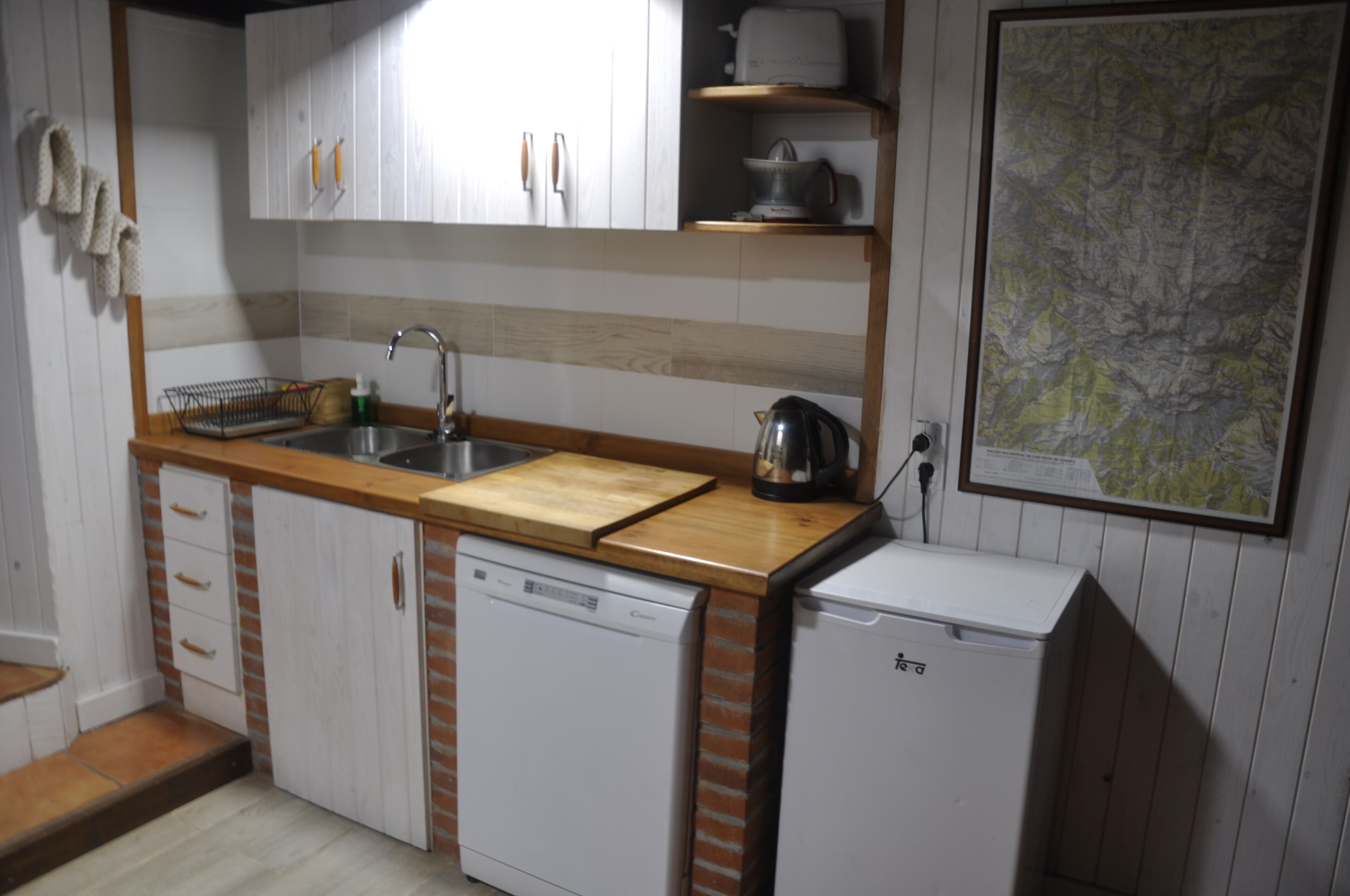 Una cocina compacta con armarios blancos, una encimera de madera, un fregadero, lavavajillas y un refrigerador pequeño.