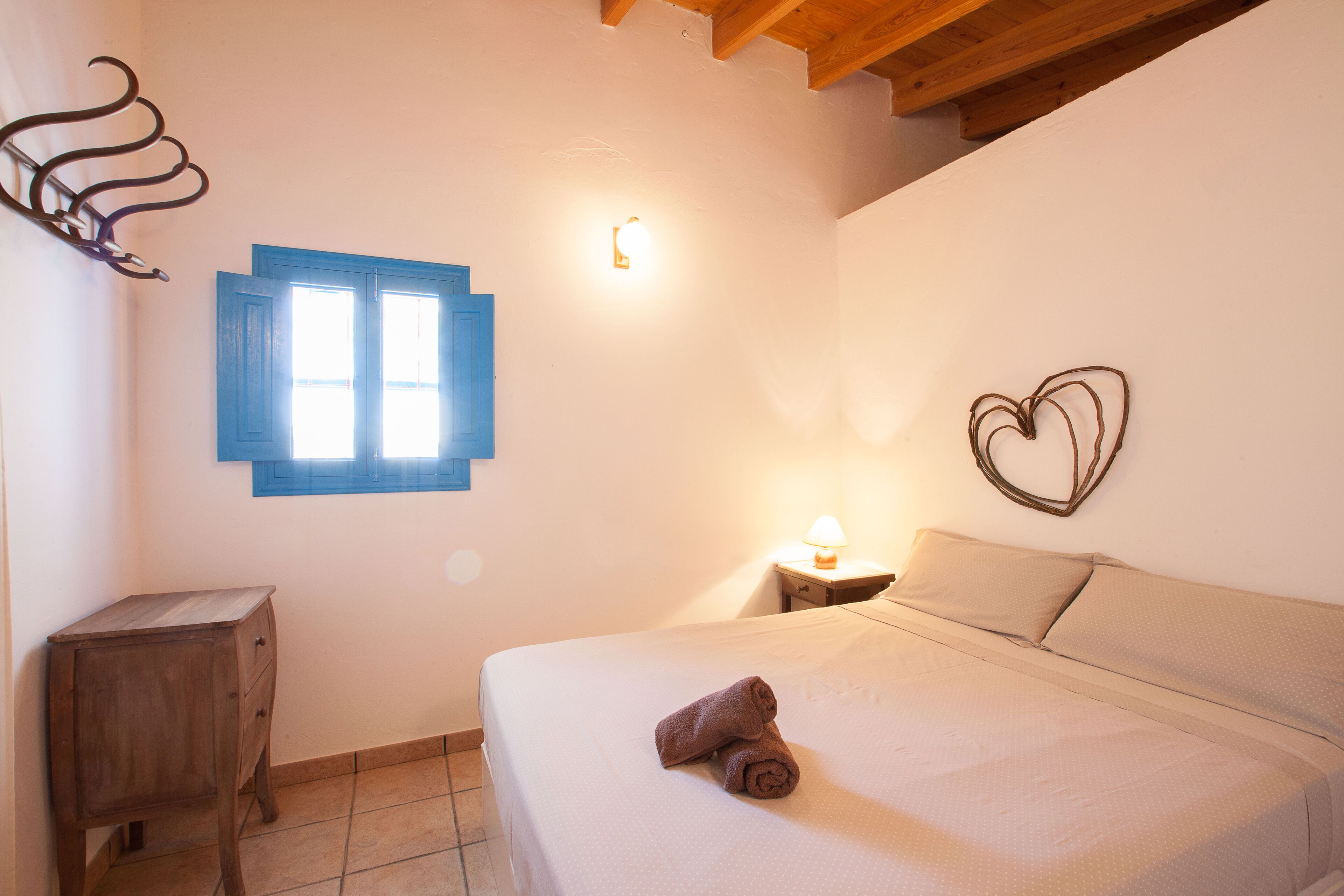 Un dormitorio con cama, mesita de noche, luz empotrada en la pared, ventana con contraventanas azules y una decoración con forma de corazón en la pared.
