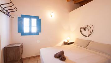 Un dormitorio con cama, mesita de noche, luz empotrada en la pared, ventana con contraventanas azules y una decoración con forma de corazón en la pared.