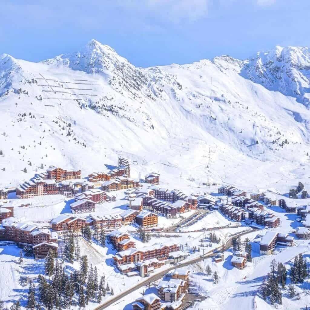 Een besneeuwde bergstad met meerdere gebouwen, waaronder hotels en mogelijk skiolatjes, omgeven door besneeuwde hellingen en bomen.