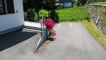 Un estacionamiento con un edificio blanco, un muro de piedra y una colina cubierta de césped con una estructura de madera al fondo.