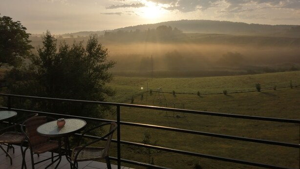 Ein Balkon mit Tisch und Stühlen mit Blick auf eine nebelverhangene Landschaft, vor der die Sonne hinter Hügeln versinkt.