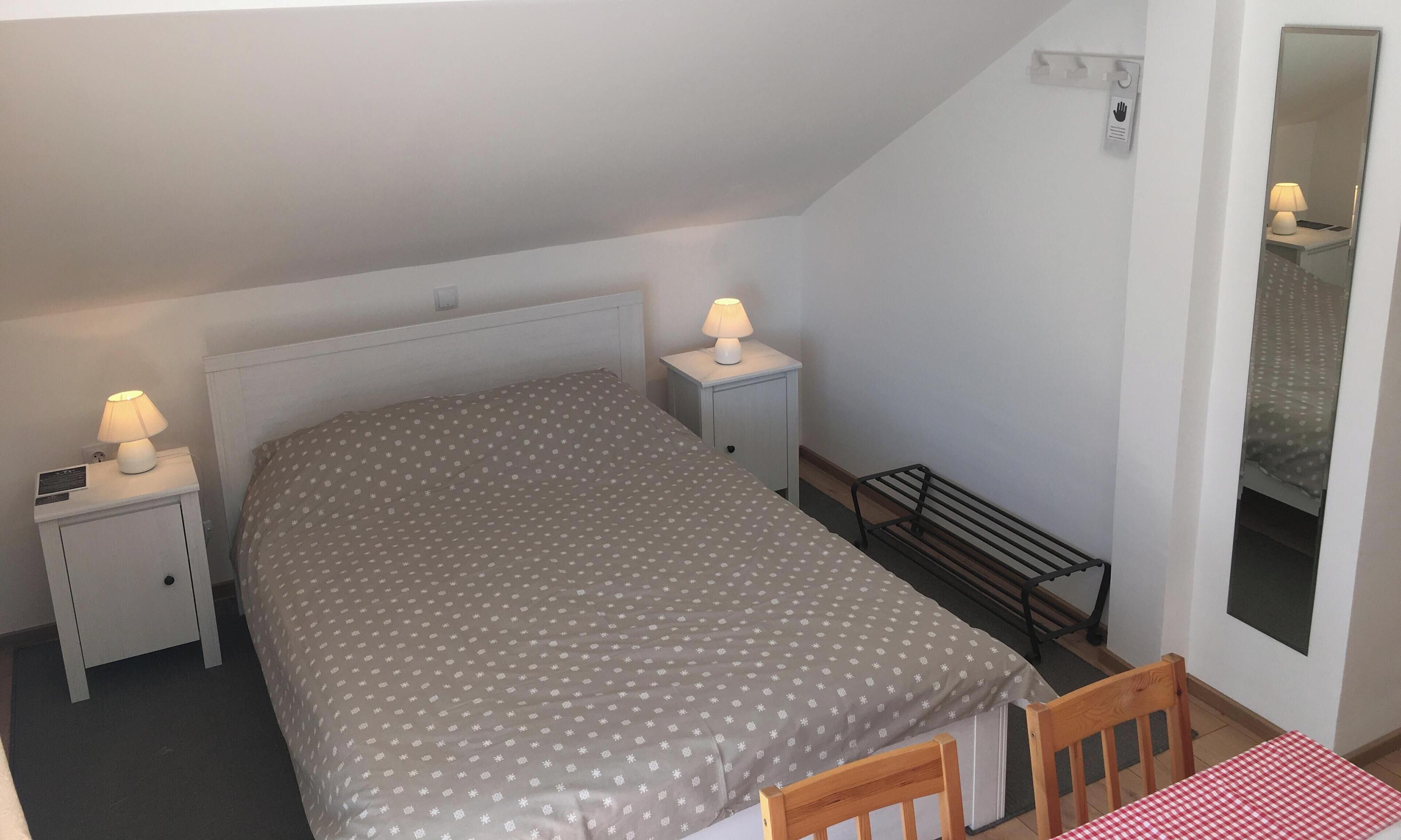 Une chambre à coucher avec un lit, deux tables de chevet avec des lampes, une chaise en bois et une petite table recouverte d’une nappe à carreaux rouges et blancs.