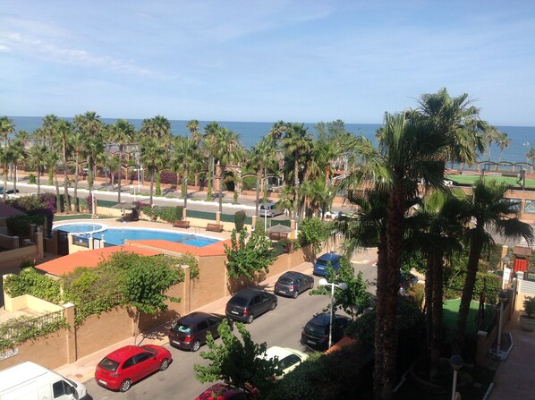A view of a parking area with cars, a swimming pool, and palm trees.