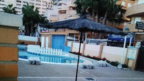 Zona de piscina con una sombrilla azul, una fuente y un edificio con un cartel que dice "BAILE ARIDO DE AGUA".