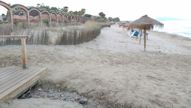 Una playa arenosa con sombrillas de paja y un camino de madera que conduce a una estructura frente al mar.