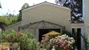 Un edificio de piedra con un patio techado, rodeado de plantas en flor y una valla blanca.