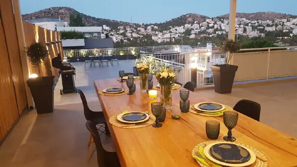 A rooftop dining area with a wooden table set for four, surrounded by chairs, with a cityscape view in the background. - Great Terrace and perfect view in Vouliagmeni (Vouliagmeni)