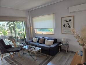 A modern living room with a grey sofa, a coffee table, and a chair. There is a window with a blind, a framed artwork on the wall, and a potted plant on a side table.