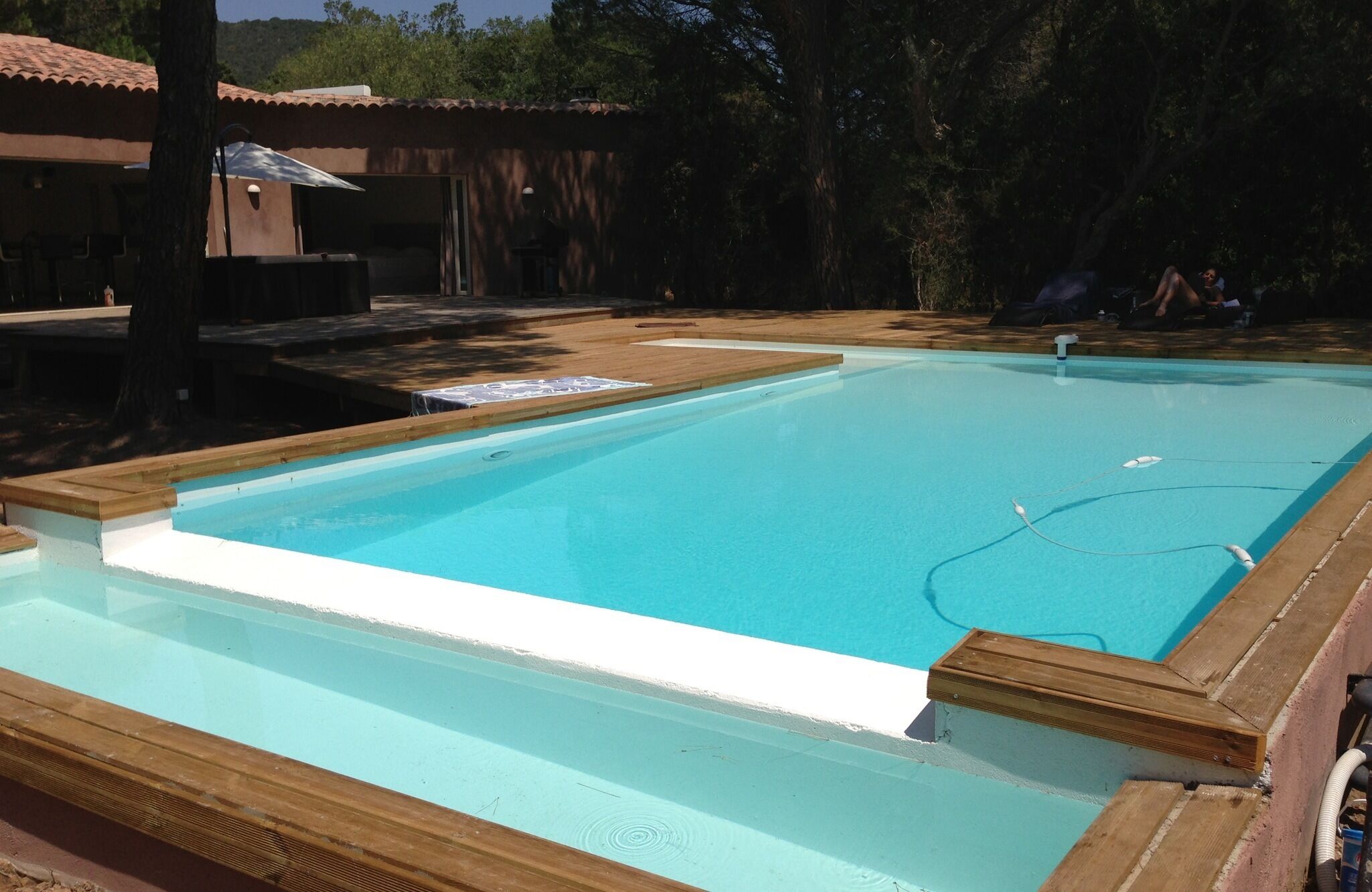 A rectangular swimming pool with a wooden deck edge, surrounded by trees and a building with a tiled roof.